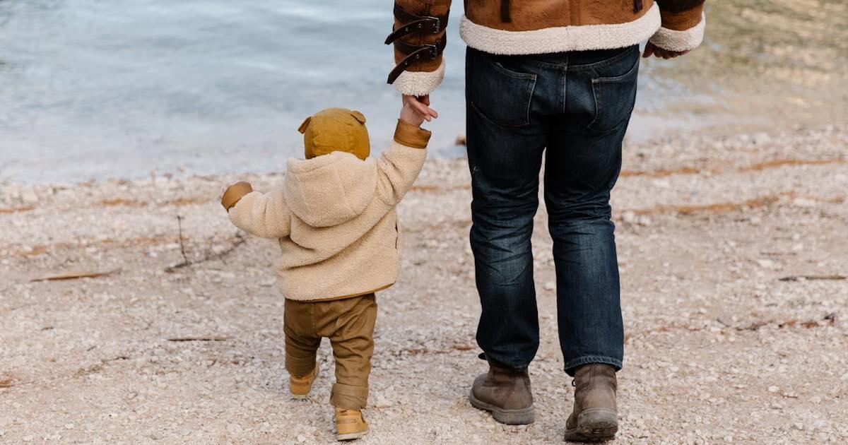 A man and child walking along the shore of a calm lake on a sunny day.