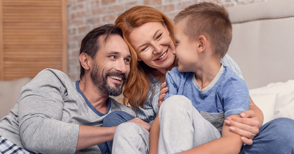 A smiling mother and father sit on a bed embracing their young son, sharing a warm and joyful moment together.