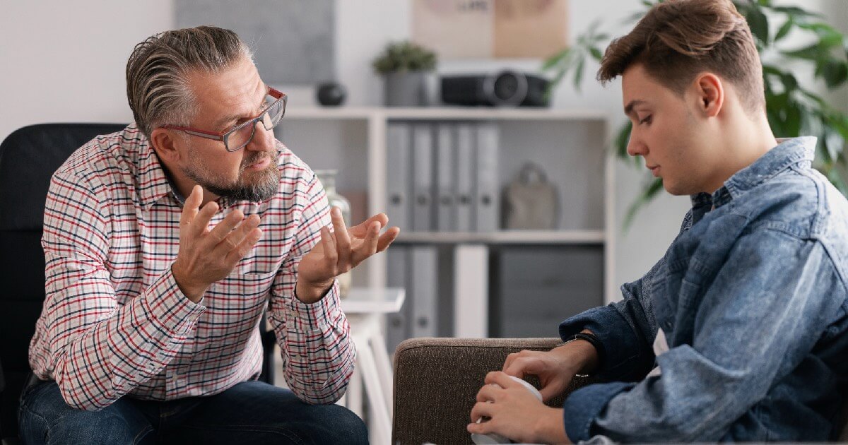 A middle-aged man gestures thoughtfully while speaking to a teenage boy who looks down, suggesting a serious one-on-one conversation.