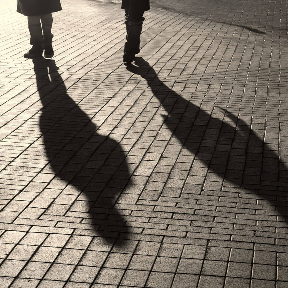 A black-and-white overhead shot of two people's feet and their long shadows cast across a textured brick walkway as they walk side by side.